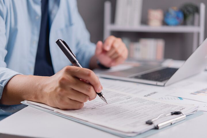 A person holding a pen behind a silver laptop writing on some sheets of paper.