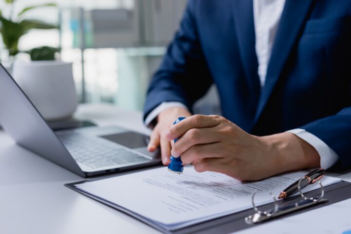 A person sits behind paperwork and a silver laptop holding a stamp.