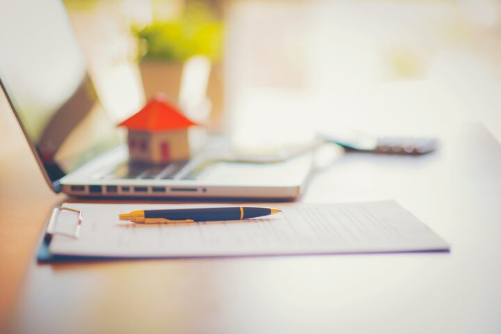 A piece of paper with a pen resting on it sits next to an open laptop with a model of a house sitting on it.